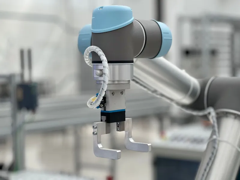 Parallelgreifer mit zwei Fingern am Flansch eines kollaborierenden Roboterarms, im Hintergrund eine unscharfe Montagelinie
