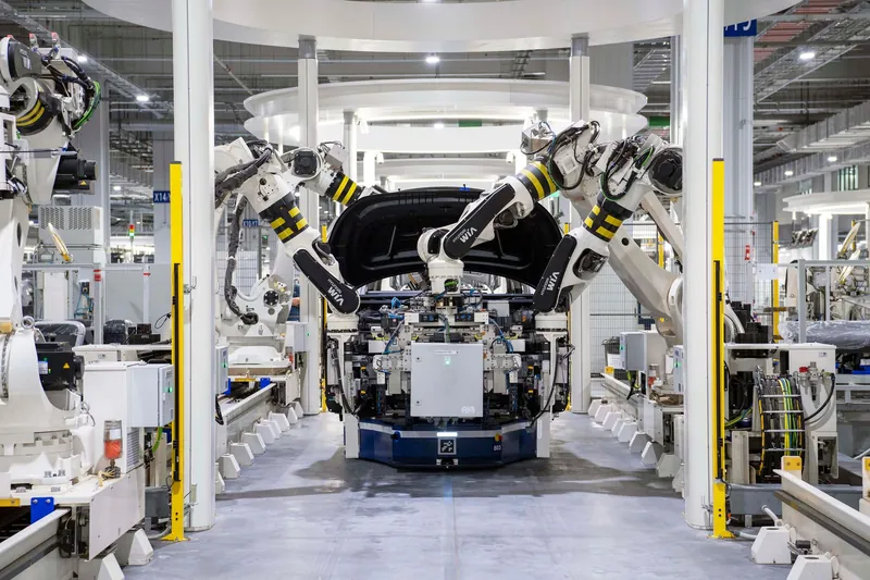 Mehrere Industrieroboter führen ein Karosserieteil über einer Fördereinheit in einer hellen Fertigungshalle zusammen